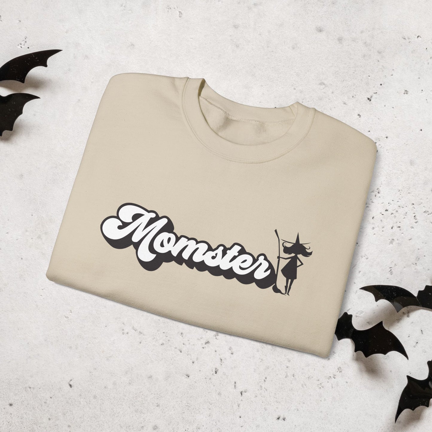Folded Beige crewneck sweatshirt with 'Momster' text and a halloween witch on a textured surface.