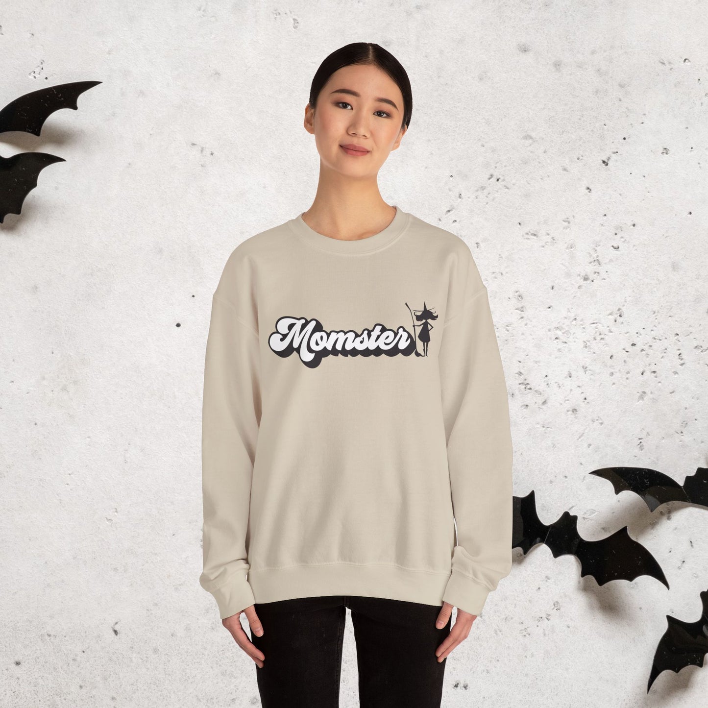 Person wearing a beige crewneck sweatshirt with 'Momster' print and Halloween witch against a textured white background with bat decorations.