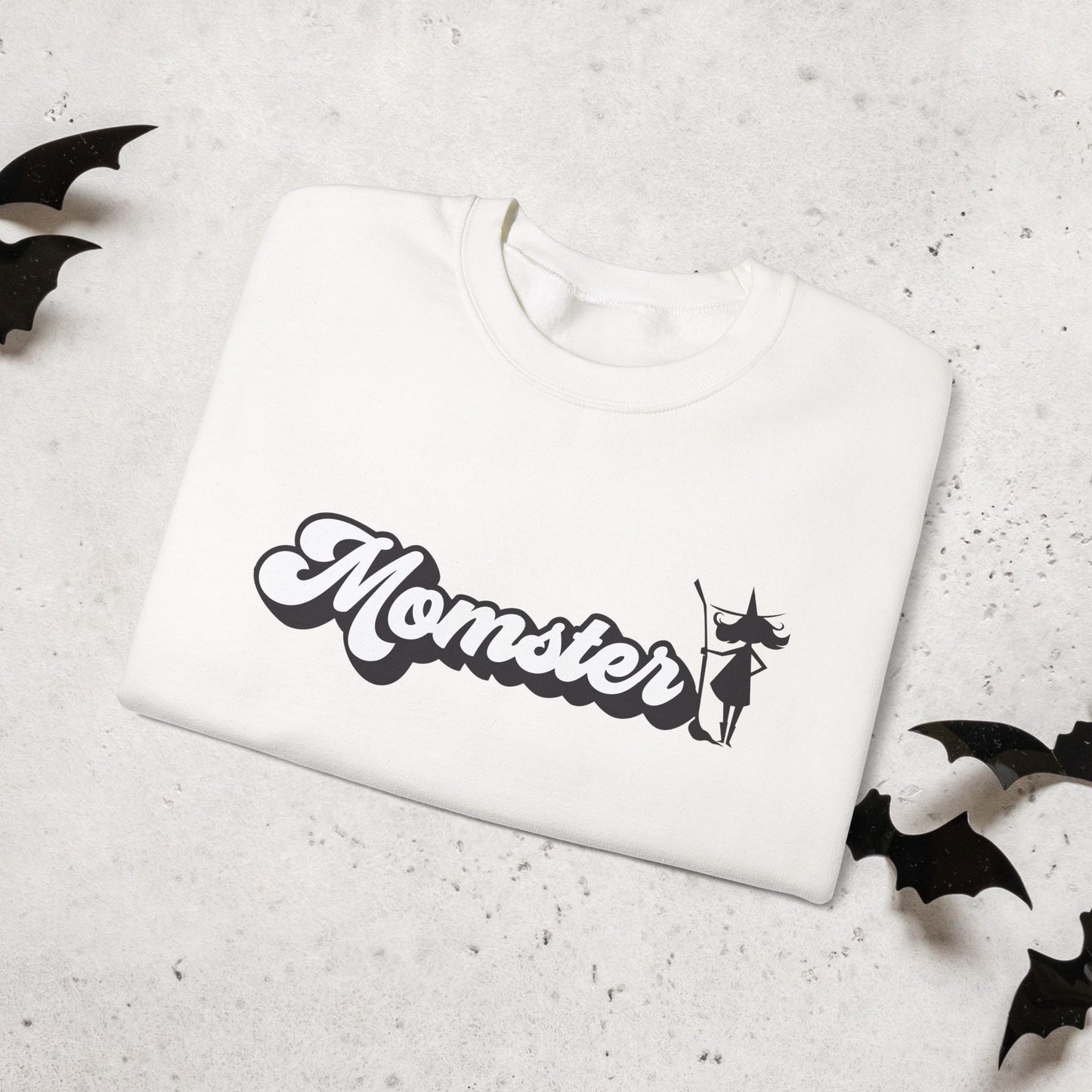 Folded White sweatshirt with 'Momster' and halloween witch graphic on a light gray surface with black bat shapes.