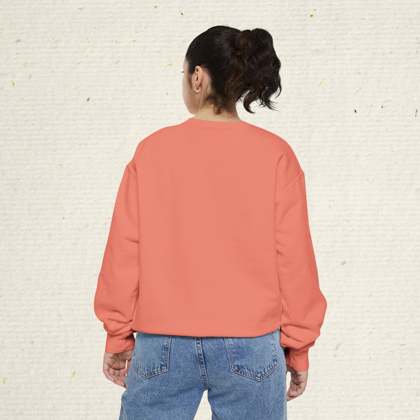 Person wearing a pink sweatshirt and blue jeans against a textured beige wall.