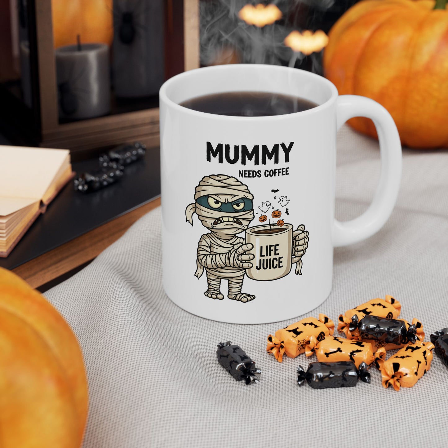 White Halloween mug with mummy design and text, surrounded by Halloween-themed items on a table.