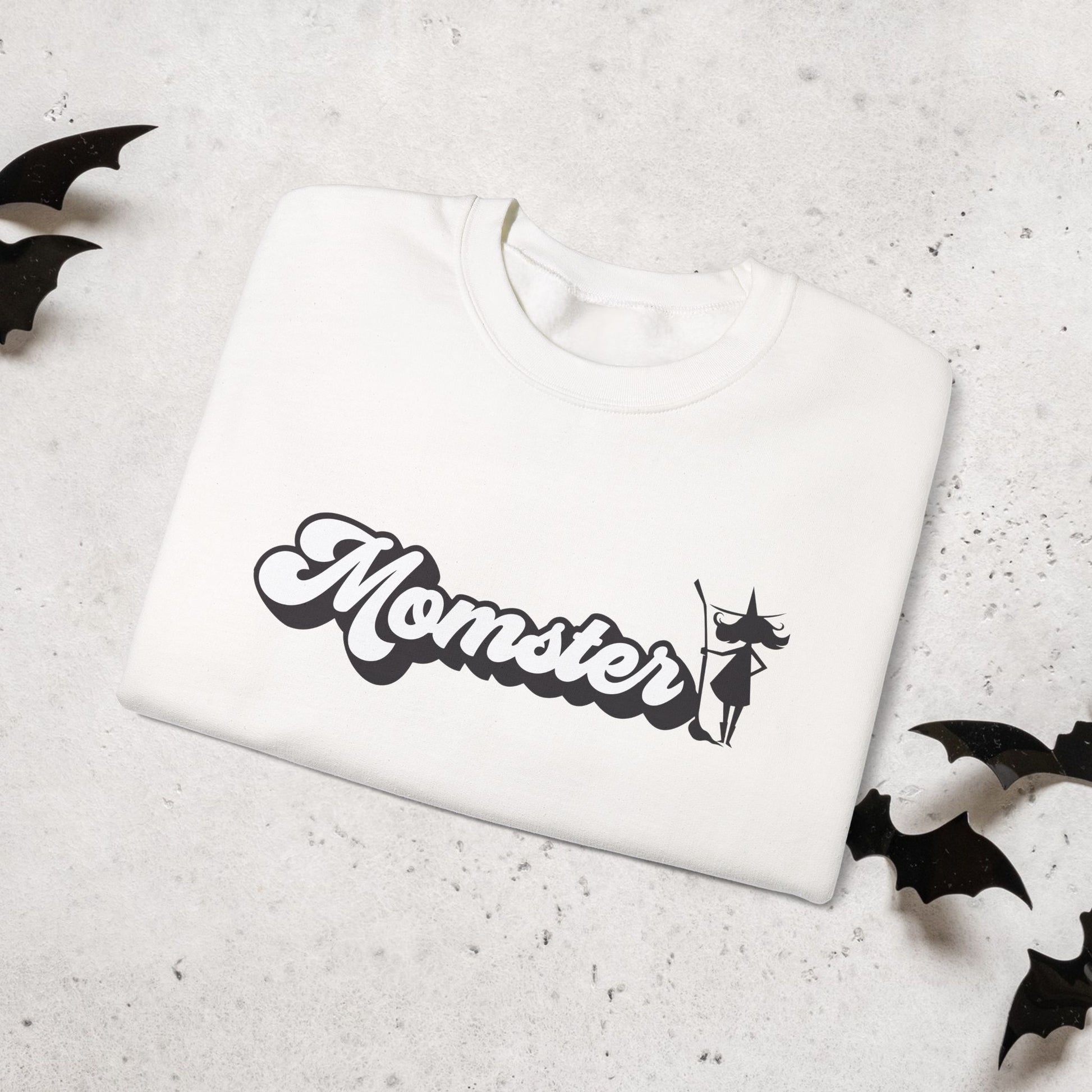 Folded White sweatshirt with 'Momster' and halloween witch graphic on a light gray surface with black bat shapes.