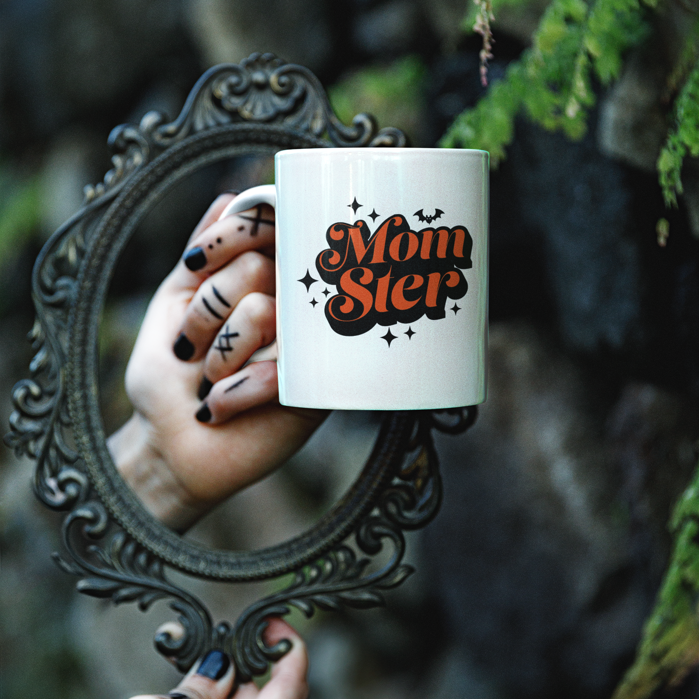 Hand holding a Halloween mug with 'Momster' text in front of a decorative mirror with a natural background